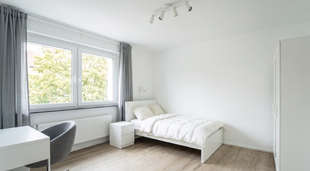 Eingerichtetes Zimmer mit Bett, Nachttisch, Kleiderschrank, Schreibtisch und Gardinen